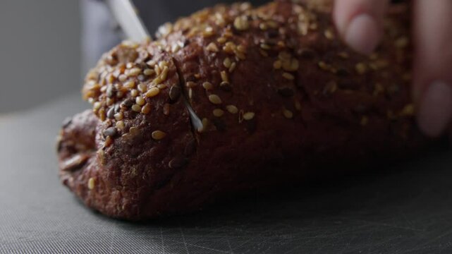 A close-up shot of a person slicing a loaf of fresh multigrain bread with a knife. The bread is covered with seeds, showcasing a rustic texture and appetizing crust.