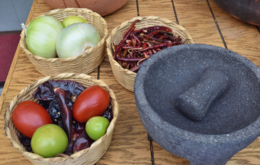 A table with a bowl of tomatoes, a bowl of green peppers, a bowl of onions, and a mortar and pestle