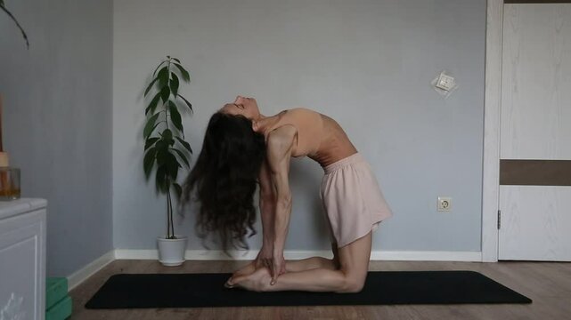 Beautiful fit atletic body woman stratching for shoulders, scapula and healthy back in bra and sporty wear doing asana camel pose on yoga mat at home interior with green flower. Ustrasana. 