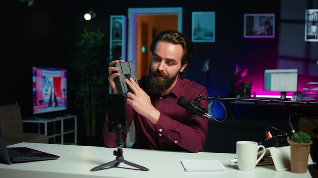Tech guru presenting V mount battery, filming technology review using cellphone on tripod and professional microphone. Caucasian vlogger showcases charging device specs, recording with phone, camera B