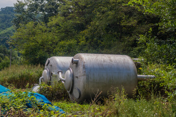 Large metal industrial tanks sit in an overgrown area, surrounded by forest greenery.