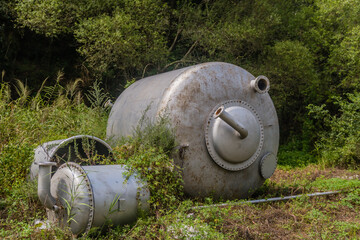 Large metal industrial tanks sit in an overgrown area, surrounded by forest greenery.