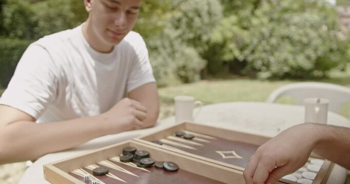 Two individuals enjoying a game of backgammon in a garden setting on a sunny day, captured up-close with game pieces in action.