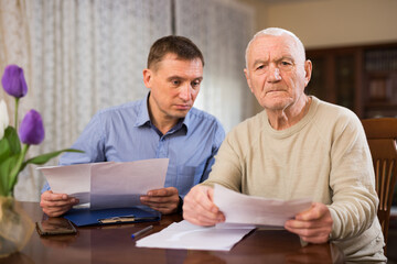 Troubled senior man discussing documents with his adult son while sitting at home table