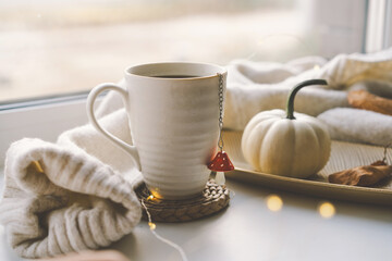 A steaming cup of tea rests on a woven coaster beside a small decorative pumpkin, with soft knit blankets and treats nearby, all illuminated by warm sunlight. Cozy autumn concept.