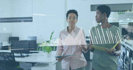 Businesswomen discussing data analysis over bar and line graphs in modern office