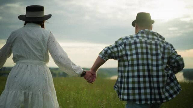 Back view of a couple holding hands and running through a grassy field at sunset. The man wears a black hat and checkered shirt, while the woman is dressed in a white dress and hat