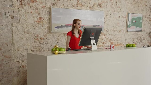 A young female receptionist stands behind a large desk and is working on a computer