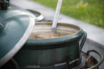 Large green metal pot with open lid and ladle serving soup outdoors on a rustic cooking setup