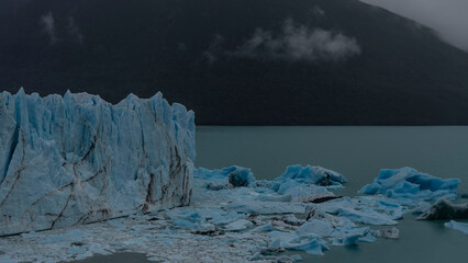 A melting glacier. A wall of cracked blue ice rises above a glacial lake. Melted ice floes float in...