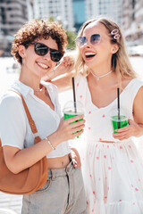 Two young beautiful smiling hipster female in trendy summer clothes. Carefree women posing outdoors. Positive models holding and drinking fresh cocktail smoothie drink in plastic cup