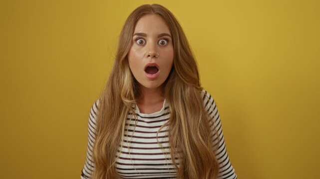 Woman shocked, young blonde wearing stripes t-shirt with amazed mouth open in disbelief, isolated over yellow background, capturing surprised and scared expressions
