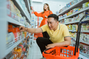 young man picking up goods on the bottom shelf by squatting while shopping with cart in minimarket