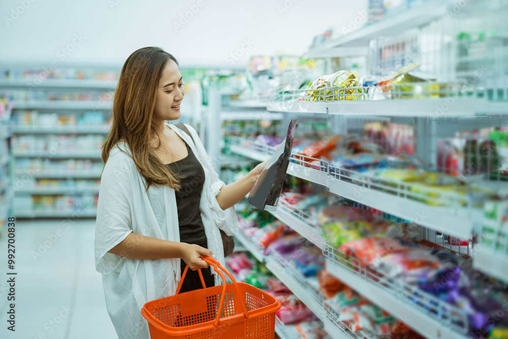 Wall mural beautiful asian woman picks up a pack of goods while shopping with a shopping cart at minimarket