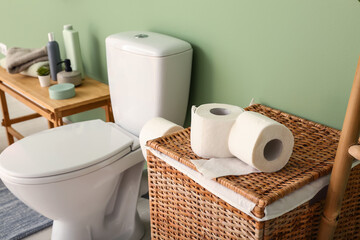 Laundry basket with paper rolls, toilet bowl and cosmetic products near color wall, closeup