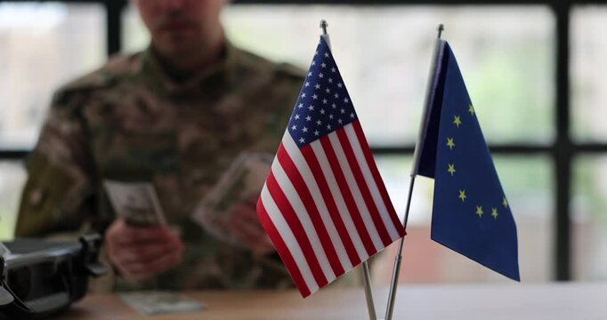 Military man in uniform sits at office desk counting money in foreign currency. Small flags of USA and European Union stand in front of army man and suitcase with money