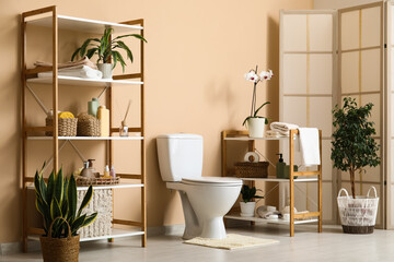 Interior of beautiful restroom with ceramic toilet bowl, houseplants and shelving unit near beige wall