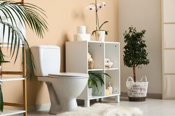 Interior of modern restroom with ceramic toilet bowl, houseplants and shelving unit near beige wall