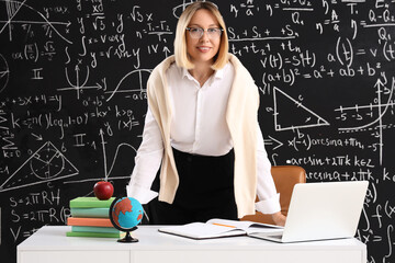 Mature female math teacher at table with laptop and globe near blackboard in classroom