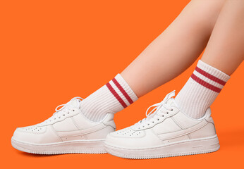 Legs of young woman in stylish white sneakers on orange background