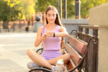 Sporty young woman sitting on bench and counting pulse after training outdoors