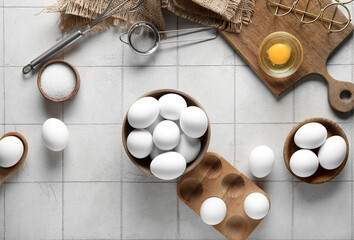 Bowls and wooden holder with raw chicken eggs on white tile background