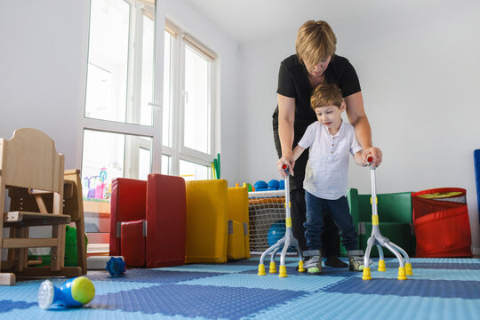 Young boy with musculoskeletal disorders uses assistive rehab canes while practicing walking with help of assistant in therapy space