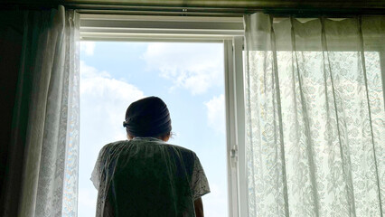 レースのカーテンがかかった窓から青空を眺める女性の後姿