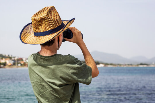 Young boy in a straw hat is looking through binoculars at the sea