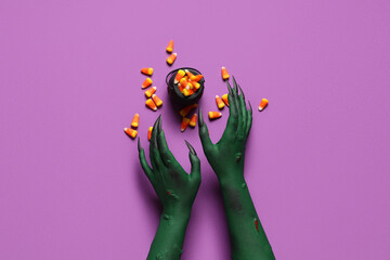 Green hands of witch with cauldron and tasty candies on purple background. Halloween celebration