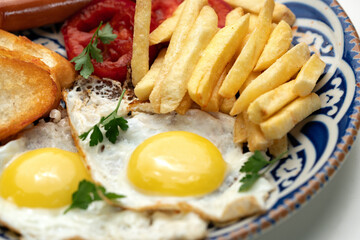 Breakfast plate with fried eggs, sausage, tomatoes, and fries served on a decorative blue plate