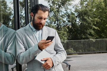 Man leaning against glass building while holding a book and checking phone, reflecting on the...