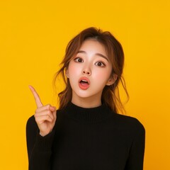 Obraz premium Surprised young woman in black sweater pointing up against bright yellow background, expressing shock or realization.