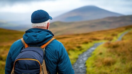 Fototapeta premium Elderly man wearing a blue jacket and cap hiking through a scenic mountain path with a backpack in a rural outdoor landscape.
