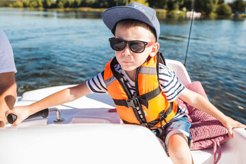 A handsome, fashionable little boy of 5 years old in a bright life jacket and sunglasses drives a...