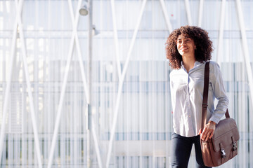 smiling female entrepreneur in smart casual outfit walking along street in megalopolis and looking away