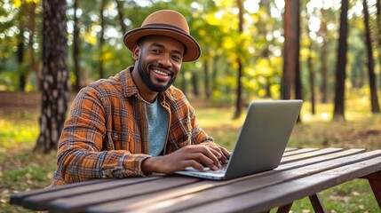 Obraz premium A smiling man is working on a laptop outdoors in a forest, wearing a stylish hat and a patterned shirt, surrounded by lush greenery.