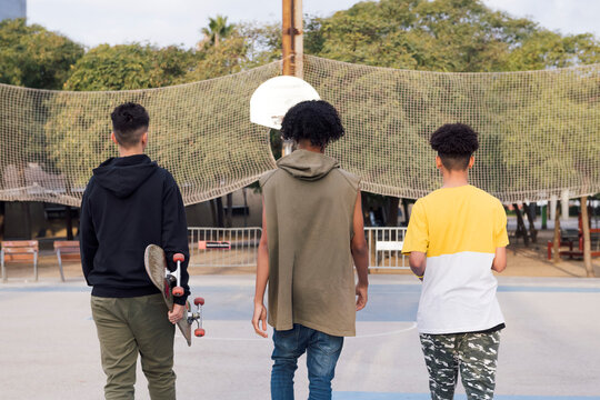 rear view of three unrecognizable multiracial male teens with skateboard walking in a sports court at park, concept of youth and urban lifestyle - Powered by Adobe