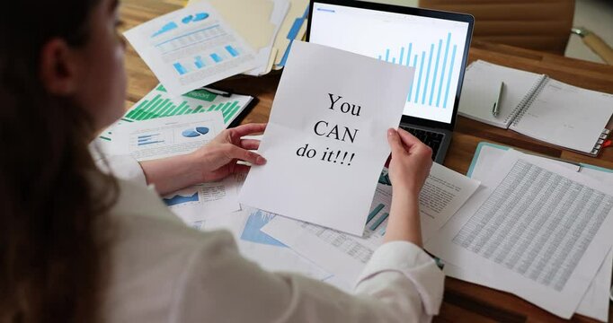 Business lady unfolds piece of paper with motivational phrase You Can Do It sitting at wooden table littered with sheets of papers with statistical data. Employee motivation