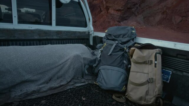 Medium no people shot of two hikers backpacks in trunk of pickup truck, parked next to stony red mountain in desert in evening, during road trip