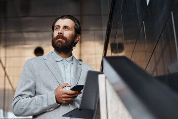 Bearded man in gray suit holding smartphone while standing in sleek, contemporary office setting with glass paneling. He has thoughtful expression on his face