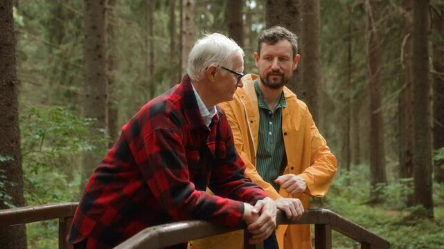 Adult son and elderly father walk together on pathway in forest, enjoying talking. Adult man supports senior people chatting and strolling. Intergenerational relationships and care for older people.