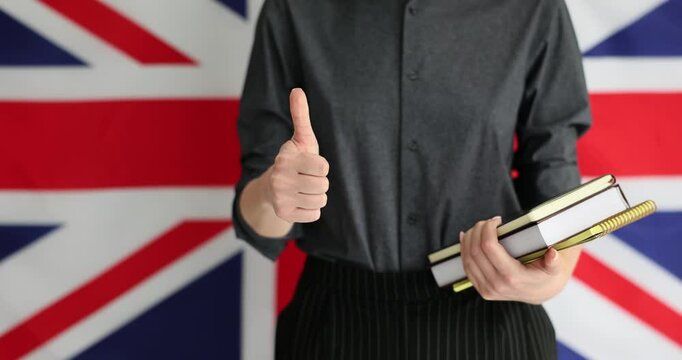 Female person with textbooks and notebooks shows gesture thumb-up after session of studying English. Brandwall of UK flag shows respect for learning English language