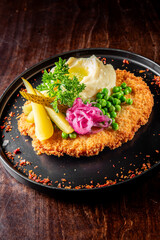A plate of breaded schnitzel served with mashed potatoes, peas, and garnish on a dark wooden table. Perfect for food and dining themes.