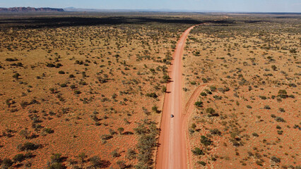 Car driving on a straight gravel road in the Australian red centre. Long sandy road with trees on the side. Drone view of outback australia. Lost in the middle of the desert. © Elsa