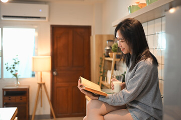 Pretty young woman with glass of milk reading book in cozy kitchen. Concept of relaxation and leisure