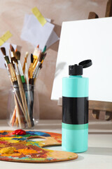 Turquoise acrylic paint in bottle, small easel, palette and brushes on wooden table indoors
