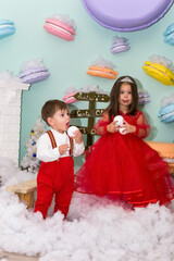 A little girl and a boy in red clothes have fun in the artificial snow by the fireplace in the studio. The girl is holding a box with gifts. Little kids in red christmas costume having fun beside a
