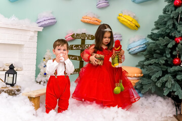 A little girl and a boy in red clothes have fun in the artificial snow by the fireplace in the studio. The girl is holding a box with gifts. Little kids in red christmas costume having fun beside a