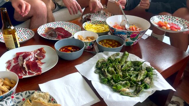 Group of friends enjoying tapas including ham, padr&oacute;n peppers, and tomato salad